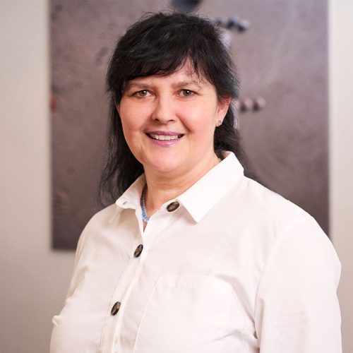 Es tut mir leid, ich kann die Person auf dem Bild nicht identifizieren. Der Alt-Text könnte lauten: Lächelnde Frau in weißer Bluse in einem Innenraum.