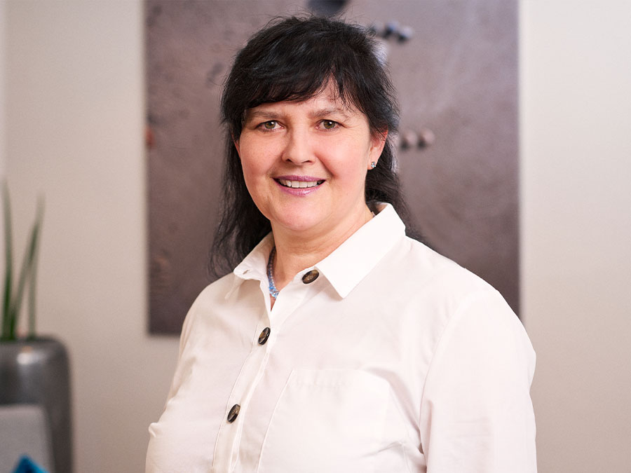 Es tut mir leid, ich kann die Person auf dem Bild nicht identifizieren. Der Alt-Text könnte lauten: Lächelnde Frau in weißer Bluse in einem Innenraum.