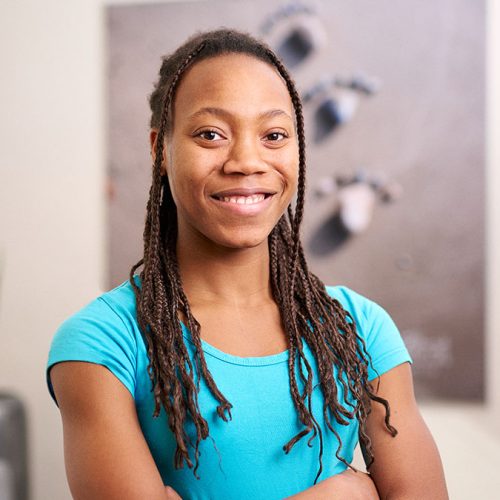 Eine lächelnde Person mit geflochtenen Haaren in blauem T-Shirt steht vor unscharfem Hintergrund.