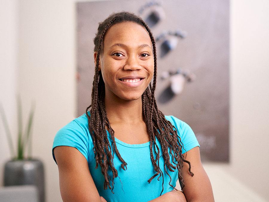 Eine lächelnde Person mit geflochtenen Haaren in blauem T-Shirt steht vor unscharfem Hintergrund.