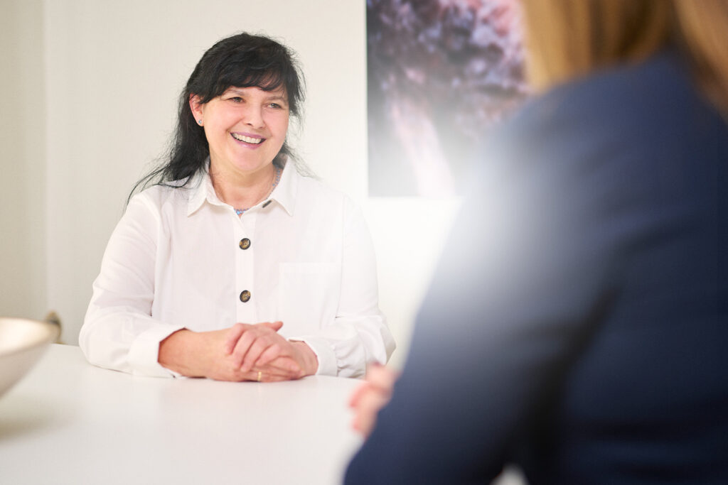 Frau im weißen Hemd lächelt während eines Gesprächs an einem Tisch, mit verschwommenem Hintergrund.