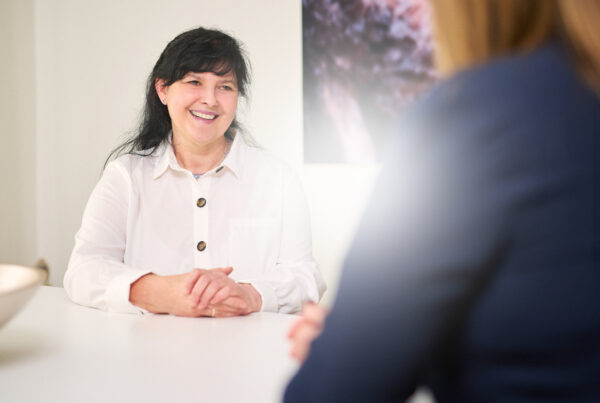 Frau im weißen Hemd lächelt während eines Gesprächs an einem Tisch, mit verschwommenem Hintergrund.