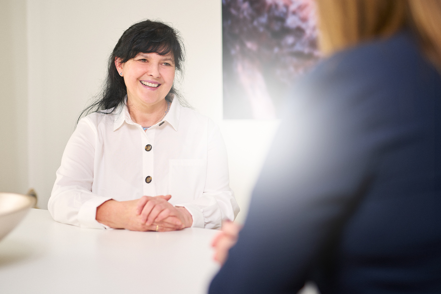 Frau im weißen Hemd lächelt während eines Gesprächs an einem Tisch, mit verschwommenem Hintergrund.